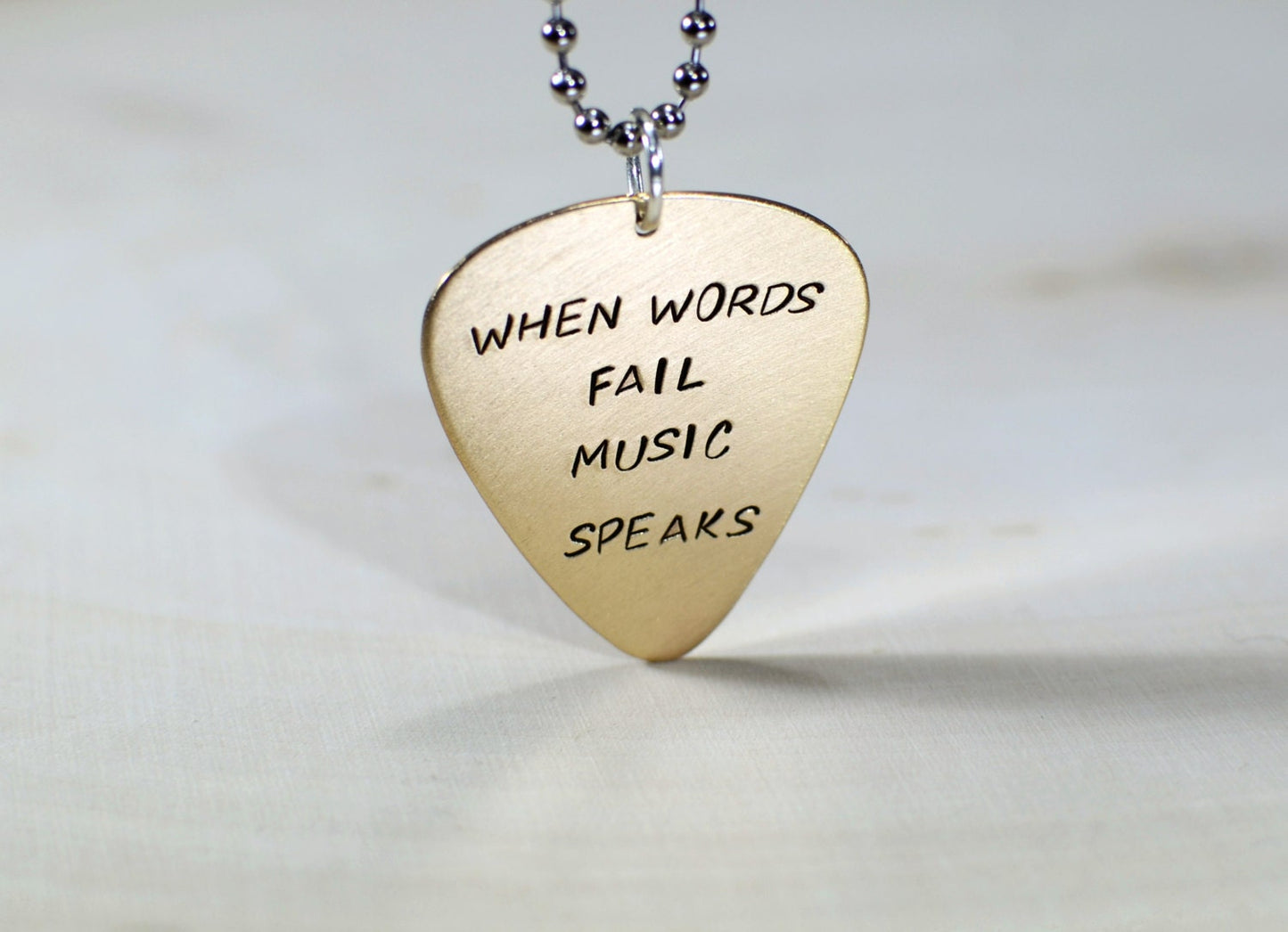 Bronze guitar pick necklace stamped with When words fail music speaks